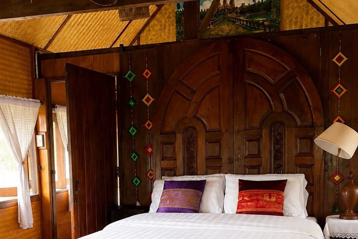 Een groot bed met houten frame, een hoofdbord met decoratieve details en uitzicht op een landschap door een raam.