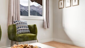 A room with a green armchair, a patterned pillow, a window with a view of mountains, and three framed pictures on the wall. - 2BR Red Cedar Lodge near Banff w/ Mountain Views (Harvie Heights)