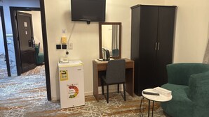 Een compacte hotelkamer met een bureau, stoel en een groene fauteuil. Een minibar en een televisie zijn aan de muur bevestigd.