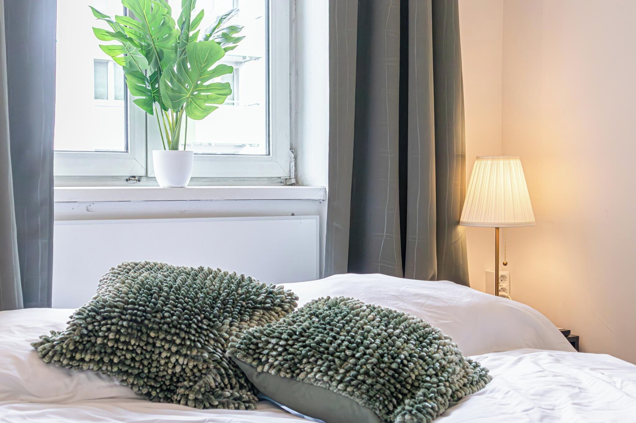 Un lit avec deux oreillers à texture, une plante en pot près d’une fenêtre et une lampe sur une table de chevet.