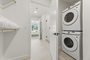 A laundry room with two stacked washing machines, a white door, and a hallway leading to a room with a bed and a window. - 1BR Suite w/ Private Balcony | Pool + Terrace + Restaurant | The Code (Austin)