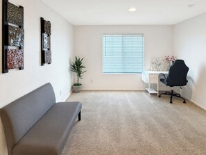 A room with a grey sofa, a desk with a chair, a window with blinds, and a potted plant. - 4BR Casa Grande Home | Game Table + Large Yard (Casa Grande)
