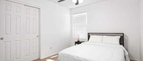 Ein Schlafzimmer mit einem Bett, einem Nachttisch, einem Deckenventilator und einem Fenster mit Jalousien.