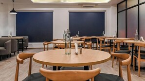 A modern dining area with round tables and wooden chairs, featuring a central bottle of water and napkins. - Townhouse D- Knowledge Park III (Greater Noida)