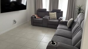 A modern living room with a grey sofa, a flat-screen TV mounted on the wall, and a ceiling fan. - Beach Holiday Home only 10mins walk to the Beach (Rainbow Beach)