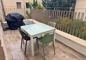 A balcony with a glass table and chairs, a barbecue grill, and a view of trees and a building. - New 2-bedroom apartment in Jerusalem for relaxing stays (Jerusalem)