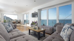 Ein geräumiger Wohnbereich mit großem Fenster zum Meer, einem an der Wand befestigten Flachbildfernseher und einer Essecke mit Tisch und Stühlen.