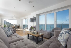 Ein geräumiger Wohnbereich mit großem Fenster zum Meer, einem an der Wand befestigten Flachbildfernseher und einer Essecke mit Tisch und Stühlen.