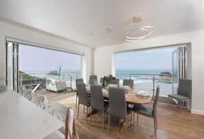 Een moderne eethoek met uitzicht op zee, met een houten tafel, grijs gestoffeerde stoelen en een glazen hekwerk.