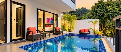 A modern pool area with a glass-enclosed bedroom, outdoor seating, and lush greenery.