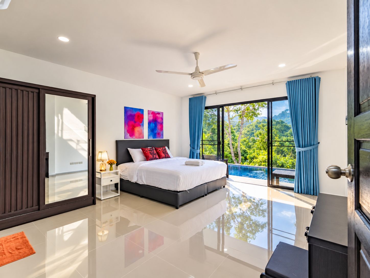 A modern bedroom with a large bed, a ceiling fan, and a view of a lush garden through floor-to-ceiling windows.