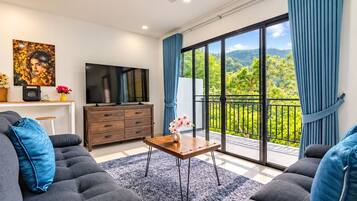 Un salon moderne doté d’un téléviseur à écran plat, d’un ensemble de canapés, d’une table basse et d’un balcon offrant une vue sur la verdure.