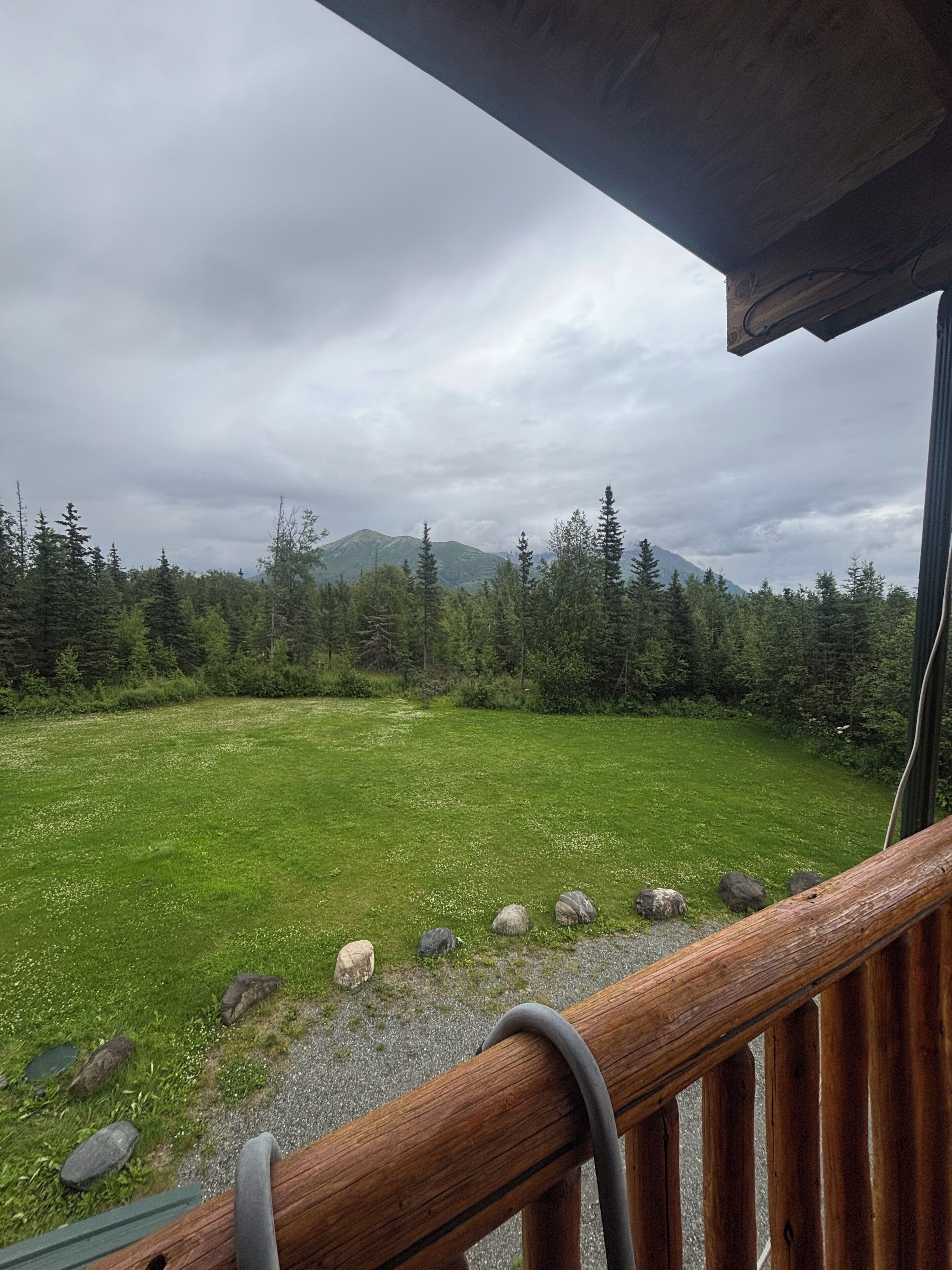 Une balustrade en bois surplombe une prairie verdoyante, avec des arbres et des montagnes visibles au loin.