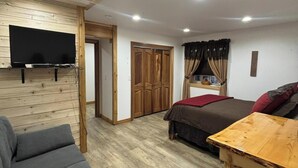 Una habitación con sofá, una mesa de madera, una cama con manta roja, un televisor montado en la pared y un armario de madera.