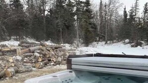 Un jacuzzi con revestimiento de madera y piedra, rodeado por un bosque y una pila de troncos.