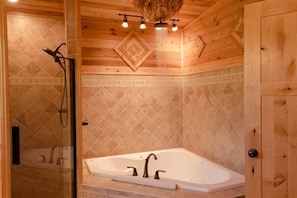 Baño de cabaña de madera con ducha envuelta en vidrio, bañera independiente y techo de madera con luces colgantes.