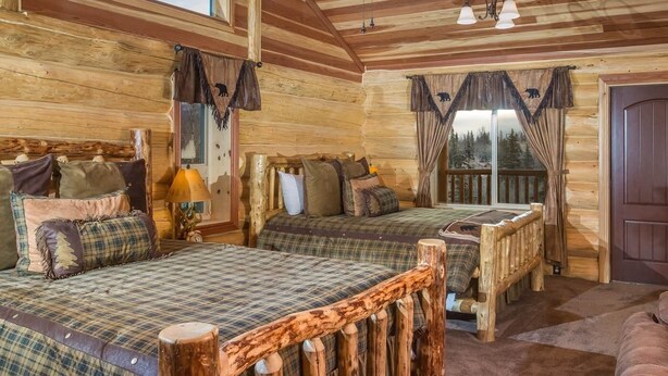 Une chambre dans un chalet en rondins, avec deux lits, une tête de lit en bois, une table de chevet et une fenêtre avec des rideaux.