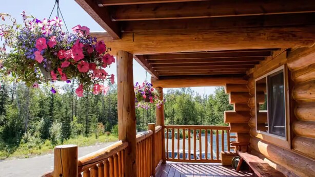 Eine Holzhütte mit einer Holzdiele, einem hängenden Blumenkorb und einem Fenster mit Blick auf einen bewaldeten Bereich.