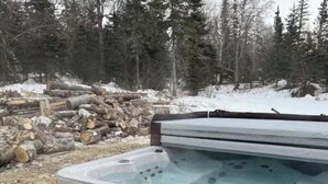 A hot tub with a wooden exterior and a stone exterior, surrounded by a forest and a pile of logs.