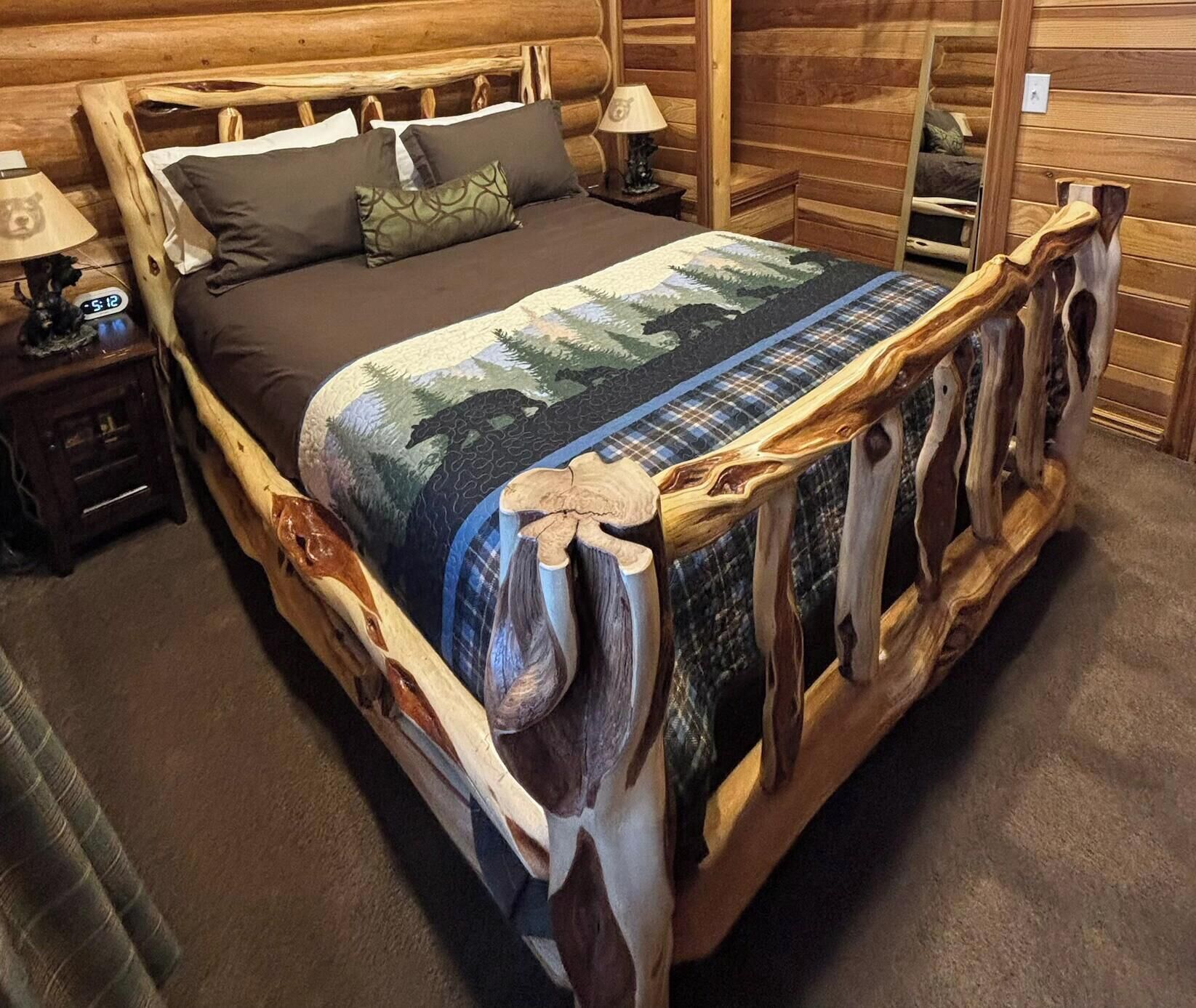 Un lit en bois avec une couvre-lit à motifs de forêt, une table de chevet avec une lampe et un miroir fixé au mur.