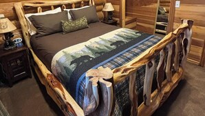 A wooden bed with a forest-themed bedspread, a nightstand with a lamp, and a wall-mounted mirror.