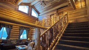 A log cabin interior with a wooden staircase, a ceiling fan, and a sofa in the living area. - Mountain View Suite~Jetted Tub~Private Balcony~3 Beds@ Alaska Knotty Pine B&B (Lazy Mountain)