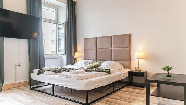 A modern bedroom with a bed, bedside tables, a TV, and a window with curtains. - Beautiful 2BR Apt Near Meidling Station (Vienna)