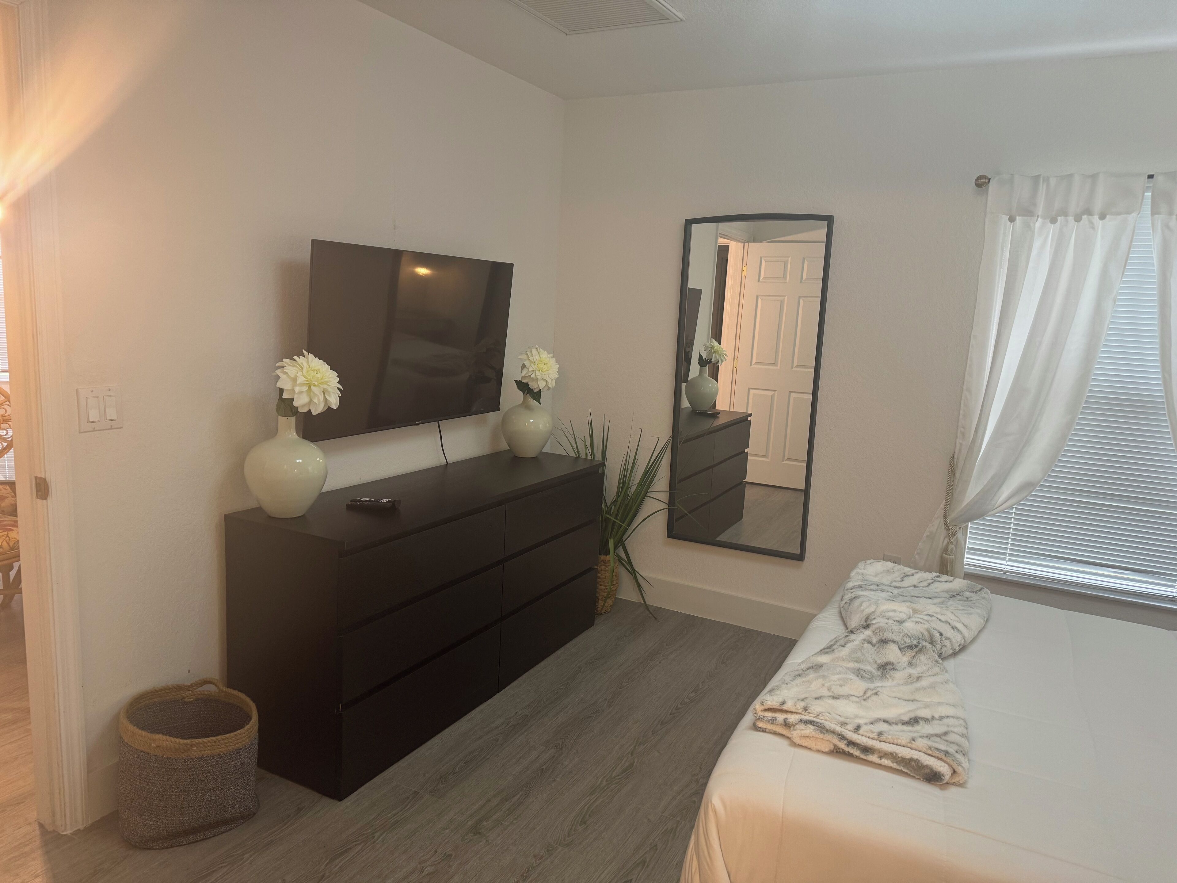 Un dormitorio con cama, cómoda, televisor, espejo y ventana con cortinas.
