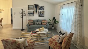 Uma sala de estar com um ventilador de teto, um sofá cinza, duas poltronas estampadas, uma mesa de centro e uma janela grande com cortinas.