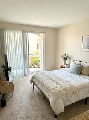 A bedroom with a large bed, a TV, a bedside table with books, and a view of a balcony with plants. - 1 Bedroom Paradise - Marina Del Rey (Marina del Rey)