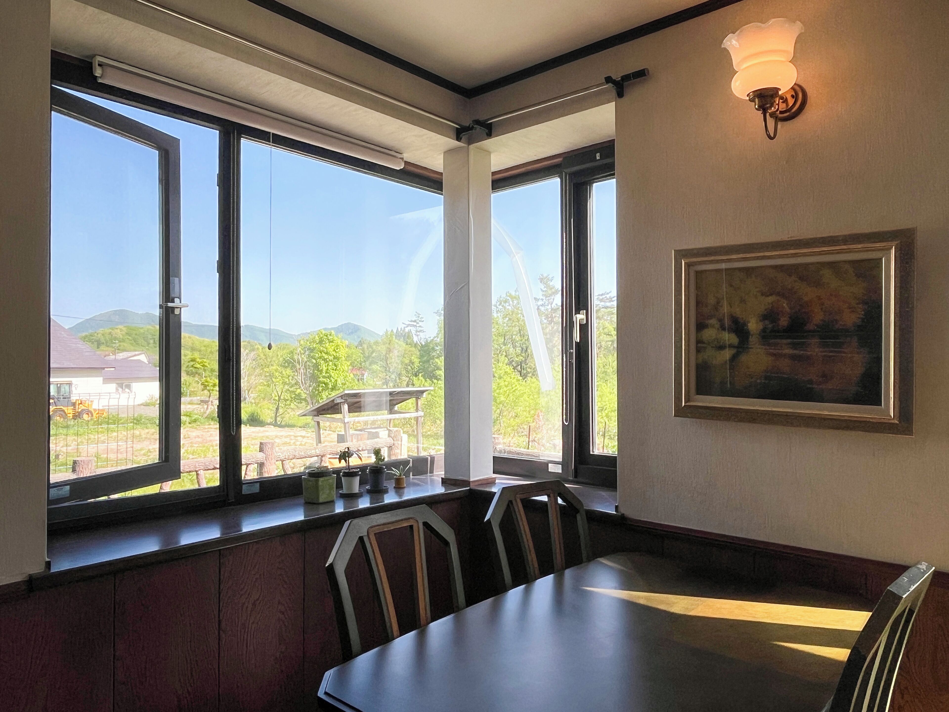 A dining area with a table and chairs, large windows with a view of greenery and buildings, and a wall-mounted light fixture.