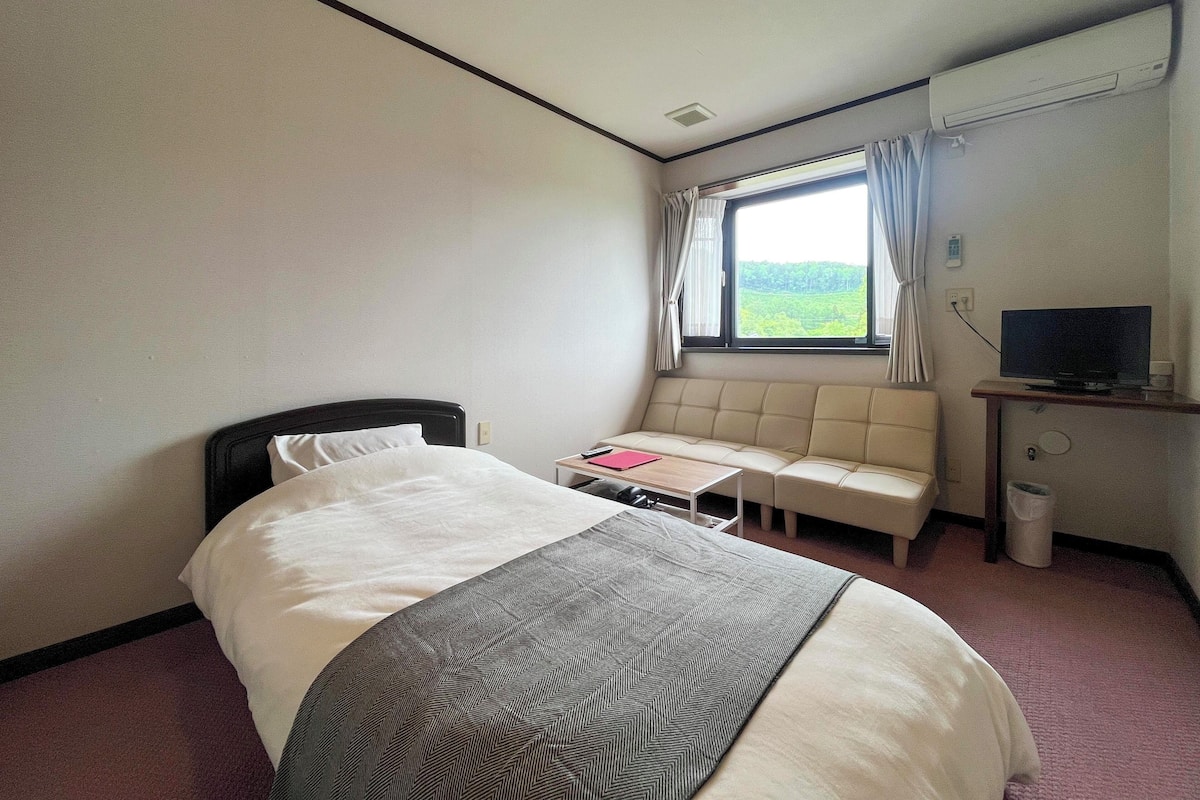 A hotel room with a bed, a small table with a red folder, a sofa, a TV, and a window with curtains.