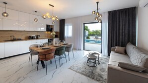 Une cuisine moderne avec des meubles blancs, un plan de travail en bois et un coin repas doté d’un sol en marbre. Le salon comprend un canapé et une table basse, ainsi qu’une grande fenêtre avec des rideaux.
