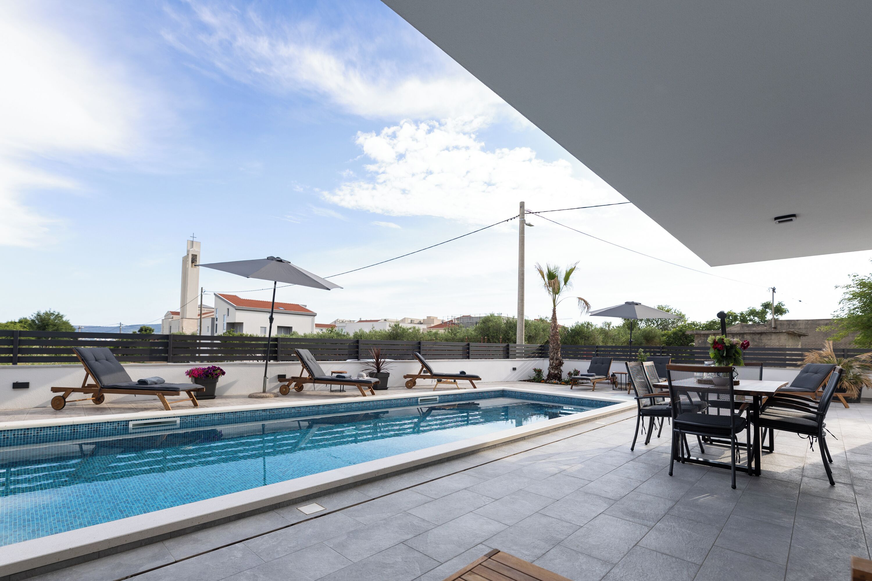 Une piscine entourée d’une clôture et de palmiers, avec des chaises longues et une table à proximité.