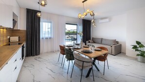 Un espace repas moderne avec une table en bois, des chaises et un lustre. On aperçoit un canapé et une plante en pot à l’arrière-plan.