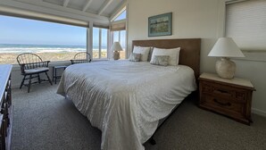 A bedroom with a large bed, two bedside lamps, a wooden nightstand, a chair, and a view of the beach. - Monterey Dunes 144 - Moss Landing Townhome - A Mile of Beachfront! (Moss Landing)
