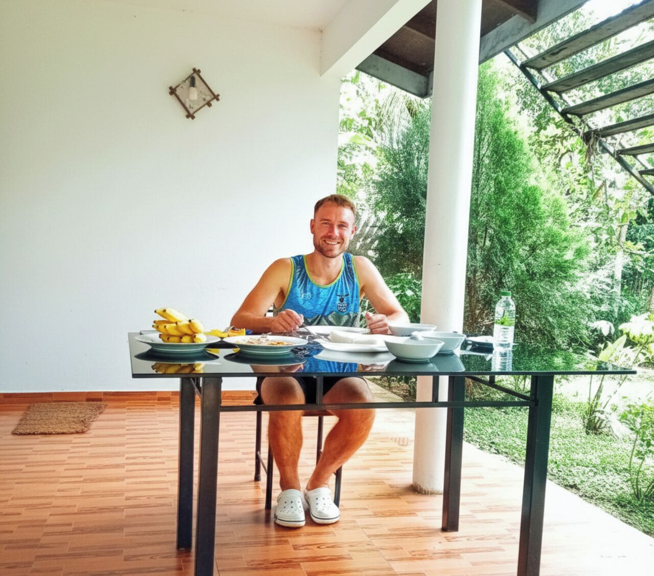Uma pessoa sentada à mesa de vidro com pratos de comida, ao ar livre, com vista para o verde.