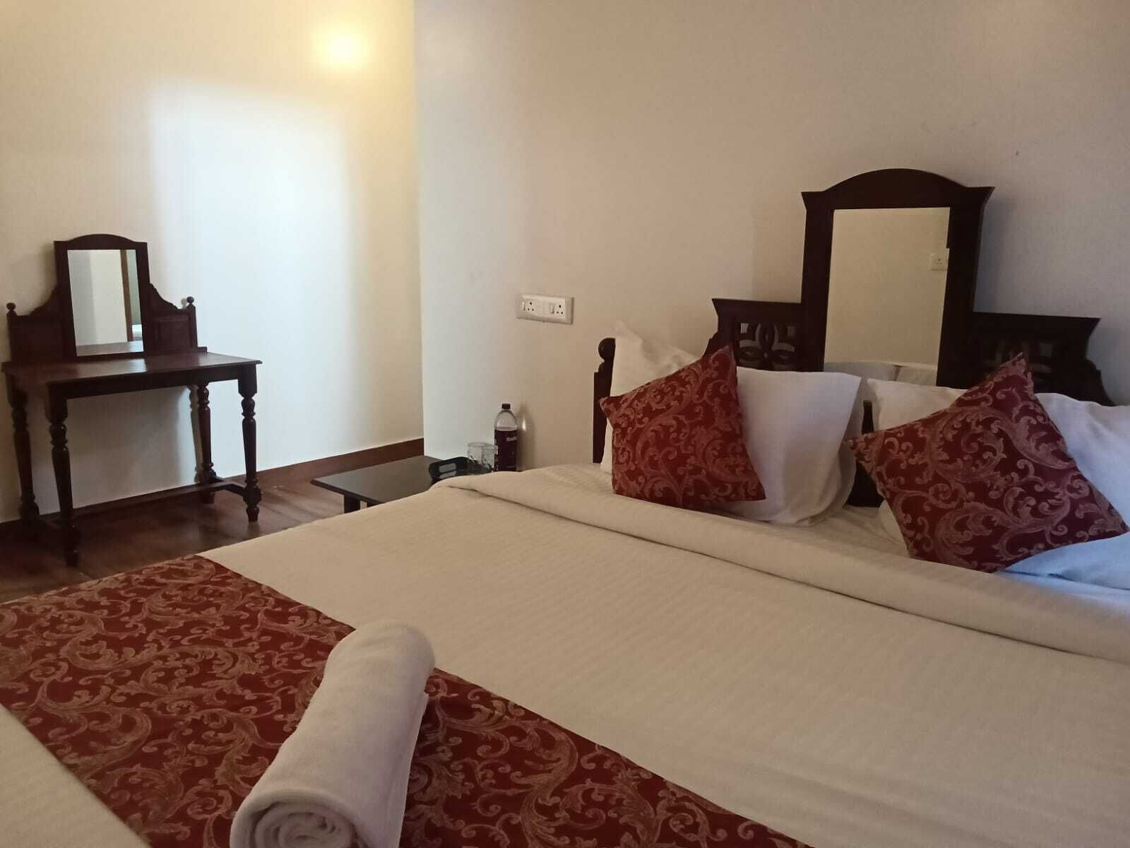 A neatly made bed with a rolled towel, a wooden nightstand, a mirror, and a wall socket.