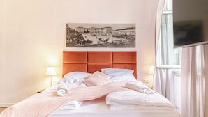 A hotel room with a bed, a headboard, a wall-mounted TV, and a painting of a historical building. - Charming 2BR Apt Near Meidling Station (Vienna)