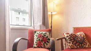 A room with two red upholstered chairs, one with a floral pillow and the other with a patterned pillow. A floor lamp is positioned between the chairs, and a window with curtains is visible in the background. - Charming 2BR Apt Near Meidling Station (Vienna)