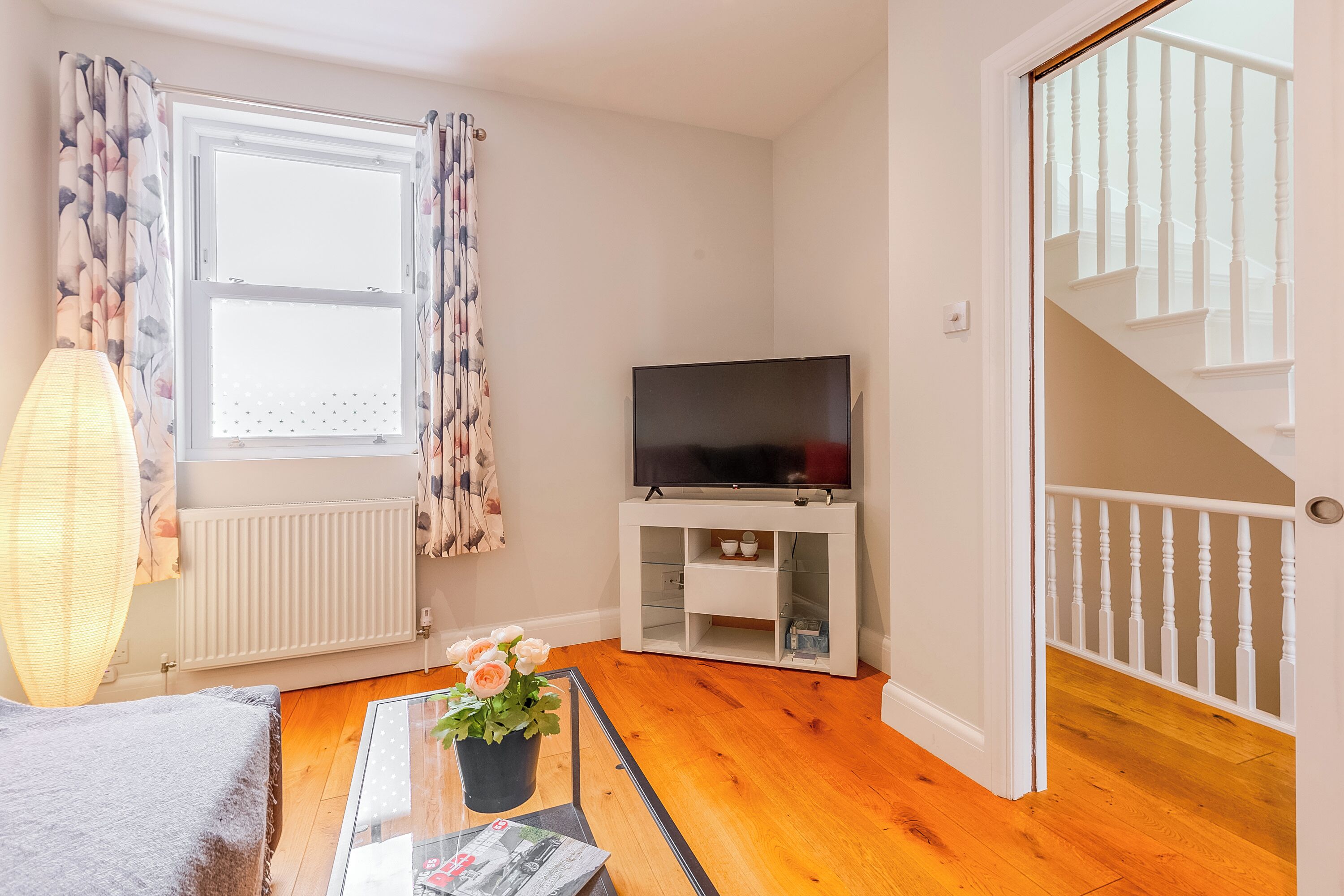 A living room with a TV, a radiator, a window with curtains, a staircase, and a coffee table with a magazine.