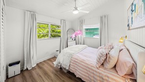 Un dormitorio con cama, ventilador de techo, ventana con cortinas, mesita de noche y lámpara de pared.