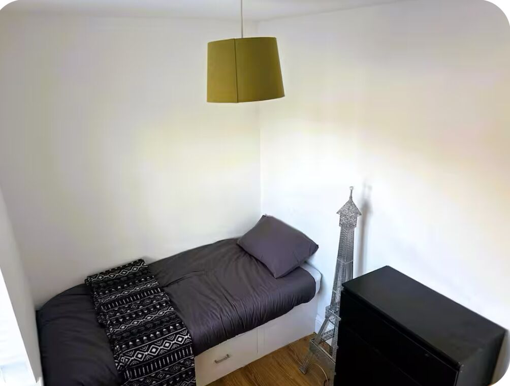 Une petite chambre avec un lit simple, une petite table, une lampe et une maquette décorative de la Tour Eiffel.