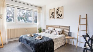 Une chambre à coucher avec un lit, un radiateur, une fenêtre avec des rideaux, une échelle et deux œuvres d’art murales.