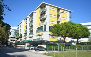 Un immeuble résidentiel à plusieurs étages, doté de balcons, d’un espace de stationnement couvert et d’un arbre au premier plan.