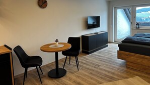 Ett modernt hotellrum med ett matbord i trä, två svarta stolar, en liten vit vas, en svart tv-apparat placerad på ett träskåp och ett stort fönster med utsikt över snötäckta kullar.