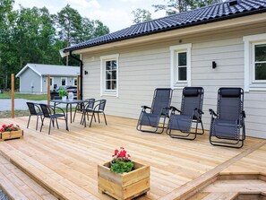 Une terrasse en bois avec une table et des chaises, un jardinier en bois orné de fleurs et une rangée de chaises longues noires.