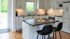 Una cocina moderna con armarios blancos, una isla central con dos taburetes altos, un horno empotrado y una ventana con vistas a la vegetación.