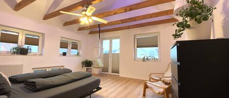 A bedroom with a bed, a ceiling fan, wooden beams, a window with blinds, a radiator, and a potted plant.
