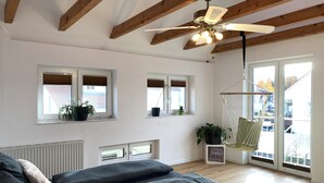 A spacious room with a high ceiling, wooden beams, a ceiling fan, and large windows with blinds.